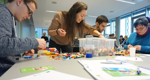 Foto einer Gruppe Studierender. Sie sitzen um einen Tisch herum und basteln mit Lego im Zuge des Workshops "Agile meets Lego".