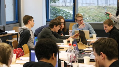 Foto von Studierenden während der Teamarbeit. Sie sitzen an einem Tisch und arbeiten an Laptops oder unterhalten sich.