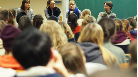 Foto von einer Gruppe von Studierenden von hinten gesehen. Vor ihnen sitzen vier Referenten und Referentinnen auf Stühlen und sprechen mit dem Publikum.