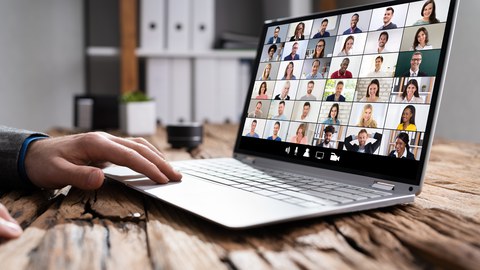 Foto eines Laptops auf einem Holztisch. Auf dem Display erkennt man in einem Raster sehr viele Gesichter, wie bei einer Videokonferenz. Eine Hand ist zu sehen, die das Touchpad des Laptops bedient.