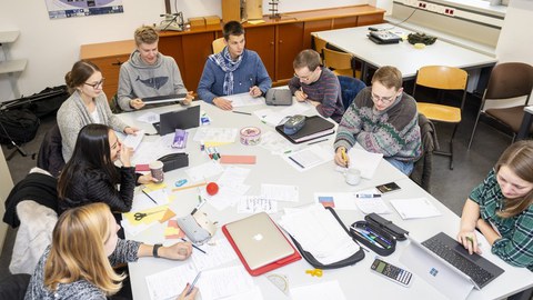 Foto von einer Gruppe Studierender. Sie sitzen um einen großen Tisch und arbeiten an Laptops oder schreiben auf Papier. Auf dem Tisch liegen viele unterschiedliche Arbeitsmaterialien wie Stifte, Blätter und Taschenrechner.