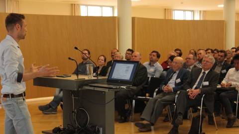 Foto eines Salles. Links im Bild steht hinter einem Pult ein Mann und hält eine Rede. Vor ihm im Raum sitzen viele Personen auf Stuhlreihen und hören ihm zu.