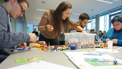 Foto einer Gruppe Studierender. Sie sitzen um einen Tisch herum und basteln mit Lego im Zuge des Workshops "Agile meets Lego".