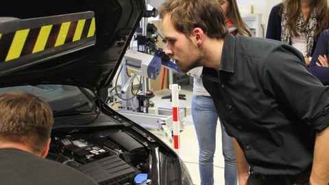 Foto eines Studenten, der unter die Motorhaube eines Autos schaut. Im Hintergrund sind zwei Studentinnen zu erkenne, die auch in das offene Auto blicken.