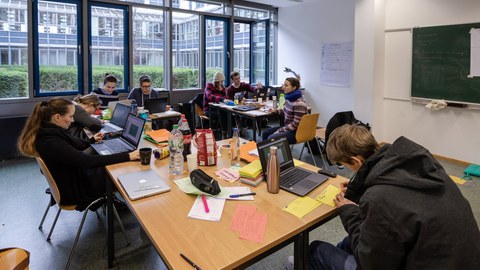 Foto von Studierenden, die an drei zusammengeschobenen Tischgruppen sitzen und an ihren Laptops arbeiten oder etwas auf Papier aufschreiben.