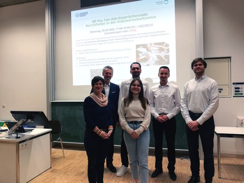 Eine Gruppe von Personen steht in einem Seminarraum vor einer Ankündigungsfolie einer Powerpoint-Präsentation und lächelt in die Kamera.