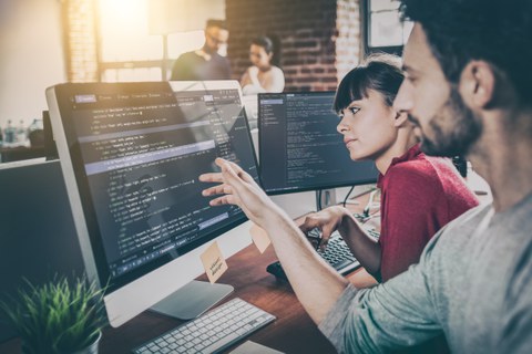 Das Foto zeigt eine Frau und einen Mann, die vor einem Computerbildschirm sitzen. Der Mann zeigt auf den Bildschirm.