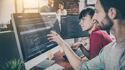 Das Foto zeigt eine Frau und einen Mann, die vor einem Computerbildschirm sitzen. Der Mann zeigt auf den Bildschirm.