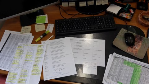Foto eines vollen Tisches, auf dem eine Tastatur, aber auch viele Klebezettel, Stifte und Blätter liegen. Am Rande des Fotos ist ein Computerbildschirm erkennbar.