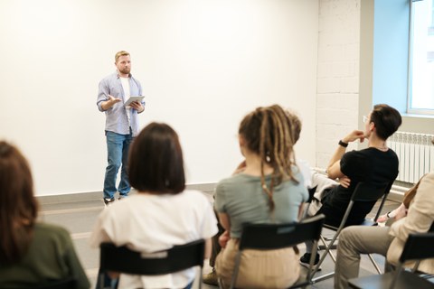 Foto eines stehenden Mannes, der vor einigen Personen auf Stühlen eine Präsentation hält. 