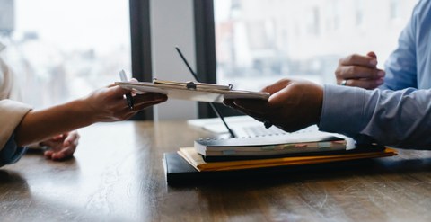 a woman's arm hands a contract to a man's arm. Under the man's arm are documents and a notebook. The people are wearing shirts/blouses. 