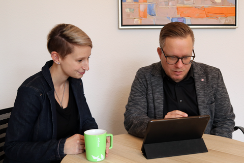 Foto einer Frau und eines Mannes, die zusammen an einem Tisch sitzen und auf ein Tablet blicken.