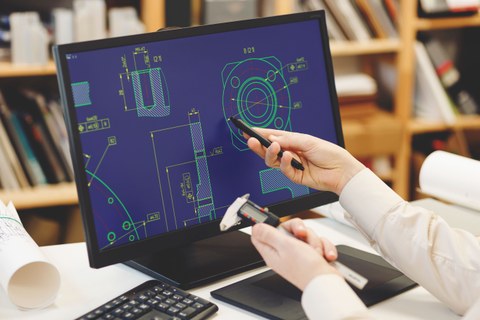 Foto eines Tisches mit Computerbildschirm, der Grafiken von Abmessungen zeigt. Eine rechte Hand deutet auf den Bildschirm und eine linke Hand hält ein Werkzeug.