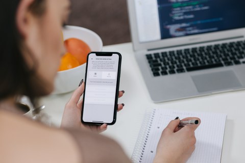 Foto einer Frau, die an einem Tisch sitzt und von hinten abgebildet ist. Sie hält ein Handy in der linken Hand und währenddessen schreibt sie mit der anderen auf einem Notizblock. Im Hintergrund ist auf dem Tisch ein Laptop erkennbar. 