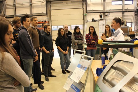 Foto von Studierenden, die in einem Halbkreis in einer Fabrikhalle stehen. Vor ihnen steht eine Frau und erklärt etwas.