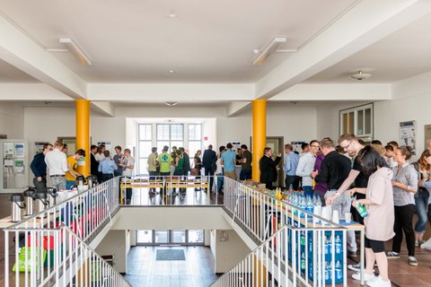 Foto einer Treppe, die in ein Foyer führt. Viele Studierende unterhalten sich dort und bedienen sich am Buffet. 