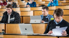 Drei Personen sind in Dokumente oder ihre Laptops vertieft und sitzen in einem Hörsaal.