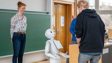 Eine Person redet mit einem humanoiden Roboter, ,eine andere schaut lächelnd zu.
