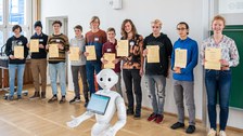 Eine Gruppe zeigt ihre Zertifikate, davor steht ein humanoider Roboter.