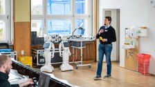 Eine Person hält einen Vortrag, im Hintergrund zwei humanoide Roboter.