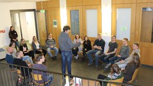 Foto während einer Kennenlernmethode. Die Studierenden sitzen in einem Stuhlkreis und ein Student steht in der Mitte des Kreises und spricht.