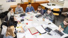 Foto von Studierenden während des Arbeitsprozesses. Die Studierenden sitzen um einen großen Tisch und diskutieren miteinander oder arbeiten an ihren Laptops. Der Tisch ist von Arbeitsmaterialien wie Blöcken, Stiften und Taschenrechnern bedeckt.