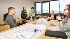 Foto einer Gruppe Studierender, die um einen großen Tisch sitzt und miteinander diskutiert. Auf dem Tisch liegt ein beschriftetes Flipchartpapier sowie Stifte.