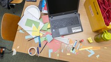 Foto eines Tisches von oben gesehen. Auf dem Tisch steht ein aufgeklappter Laptop und es liegen viele Arbeitsmaterialien wie bunte Blätter, Stifte, Kreppband, Lineale, Kleber sowie ein Taschenrechner daneben.