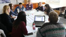Foto einer Gruppe Studierender, die um einen großen Tisch sitzt und miteinander diskutiert. Auf dem Tisch liegt ein beschriftetes Flipchartpapier sowie Stifte und weitere Blöcke und ein aufgeklappter Laptop.