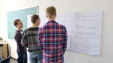Foto von drei Studenten, die drei beschriftete Flipchartpapiere betrachten, die vor ihnen an der Wand hängen.