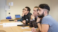 Foto der Profirunde. Die Profis sitzen in einer Reihe an einem langen Tisch und hören jemandem zu, der sich außerhalb des Fotos befindet.