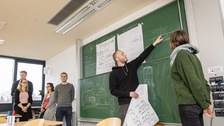 Foto einer Gruppe Studierender, die vor einer beschrifteten Tafel steht. Ein Student zeigt auf die Tafel und erklärt etwas.