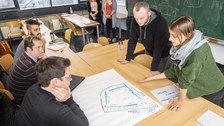 Foto von der Profirunde. Die Profis sitzen in einer Reihe an einem langen Tisch und betrachten ein beschriftetes Flipchartpapier, das vor ihnen liegt. Auf der anderen Seite des Tisches stehen zwei Studierende und erklären etwas.