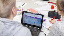 Foto von zwei Studierenden, die an einem Tisch sitzen und arbeiten. Ein Student arbeitet an einem Laptop und die Studentin rechts neben ihm schreibt mit einem Stift auf einem Block.