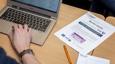 Foto eines Tisches von schräg oben betrachtet. Auf ihm liegen ein Block, ein Stift und ein Zettel mit BeING Inside Schriftzug. Weiter steht ein aufgeklappter Laptop auf dem Tisch und man erkennt eine Hand, die auf dem Laptop das Touchpad bedient.