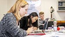 Foto von drei Studierenden, die an einem Tisch sitzen und an ihren Laptops arbeiten.