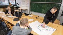 Foto eines Seminarraumes, in dem sich vier Studierende befinden. Zwei Studenten stehen sich an einem Tisch gegenüber und betrachten ein beschriftetes Flipchartpapier, das vor ihnen auf dem Tisch liegt.