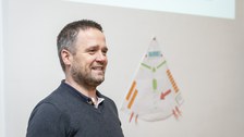 Foto eines Mannes, der etwas erklärt und der vor einer Wand steht auf der eine große Skizze befestigt ist.