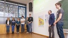 Foto einer Studierendengruppe bei der Abschlusspräsentation. Die Studierenden stehen in einer Reihe vor einer Wand. Über ihnen wird eine Präsentation an die Wand projiziert. Neben ihnen an der Wand hängen zwei beschriftete Flipchartpapiere.
