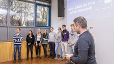 Foto einer Studierendengruppe bei der Abschlusspräsentation. Die Studierenden stehen in einer Reihe vor einer Wand. Über ihnen wird eine Präsentation an die Wand projiziert mit dem Text: "Danke für Ihre Aufmerksamkeit".