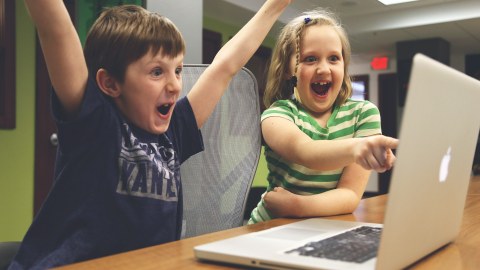 Auf dem Foto sitzen ein Junge und ein Mädchen vor einem Laptop und jubeln und freuen sich.