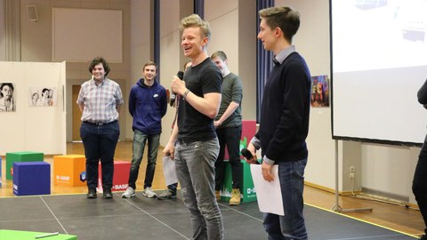 Foto von zwei Studenten, die einen Vortrag halten. Im Hintergrund stehen weitere Teammitglieder und schauen zu. An die Wand ist eine Präsentation geworfen.