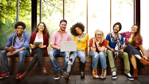 Auf dem Foto sieht man eine Gruppe junger Personen, die in einer Reihe am Fenster auf einer Bank sitzen. Die Personen lächeln und einige haben Laptops auf dem Schoß.