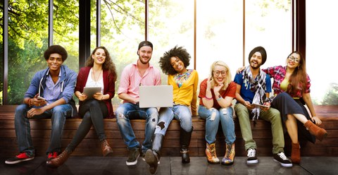 Auf dem Foto sieht man eine Gruppe junger Personen, die in einer Reihe am Fenster auf einer Bank sitzen. Die Personen lächeln und einige haben Laptops auf dem Schoß.