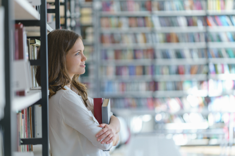 Foto einer Studentin in einer Bibliothek. Sie lehnt mit dem Rücken an einem Bücherregal und hält zwei Bücher in den verschränkten Armen. 