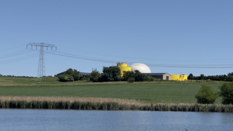 Foto: Biogasanlage  mit Freileitungsmast und Feld sowie See im Vordergrund 