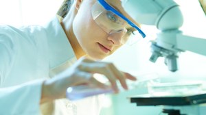Das Foto zeigt eine Studentin mit Laborkittel und Schutzbrille. Sie steht vor einem Mikroskop. Mit einem Reagenzglas gibt sie gerade eine Flüssigkeit in eine Petrischale.