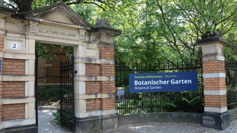Foto. Geöffnetes Tor, rechts daneben Schild mit Aufschrift: "Herzlich Willkommen! Botanischer Garten"