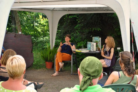 Foto: Solvejg Nitzke sitzt mit der Moderatorin Lilith Diringer an einem Tisch mit Büchern unter einem Pavillon. Vier Zuhörer:innen sehen sie an.