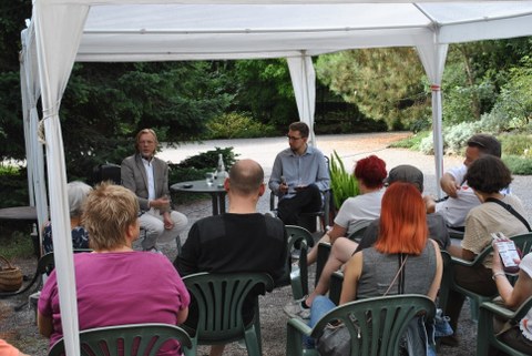 Die Fotografie zeigt an einem Tisch sitzend den Referenten Torsten Tonn und den Moderator Maurice Vetter sowie das Publikum bei einer Veranstaltung im Freien.